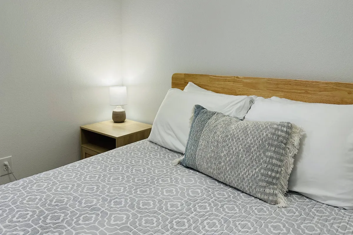 A cozy, peaceful bedroom with a bed adorned with a decorative pillow and a bedside table topped with a lamp. The room is bathed in natural light, suggesting a comfortable space for relaxing or enjoying quiet time.
