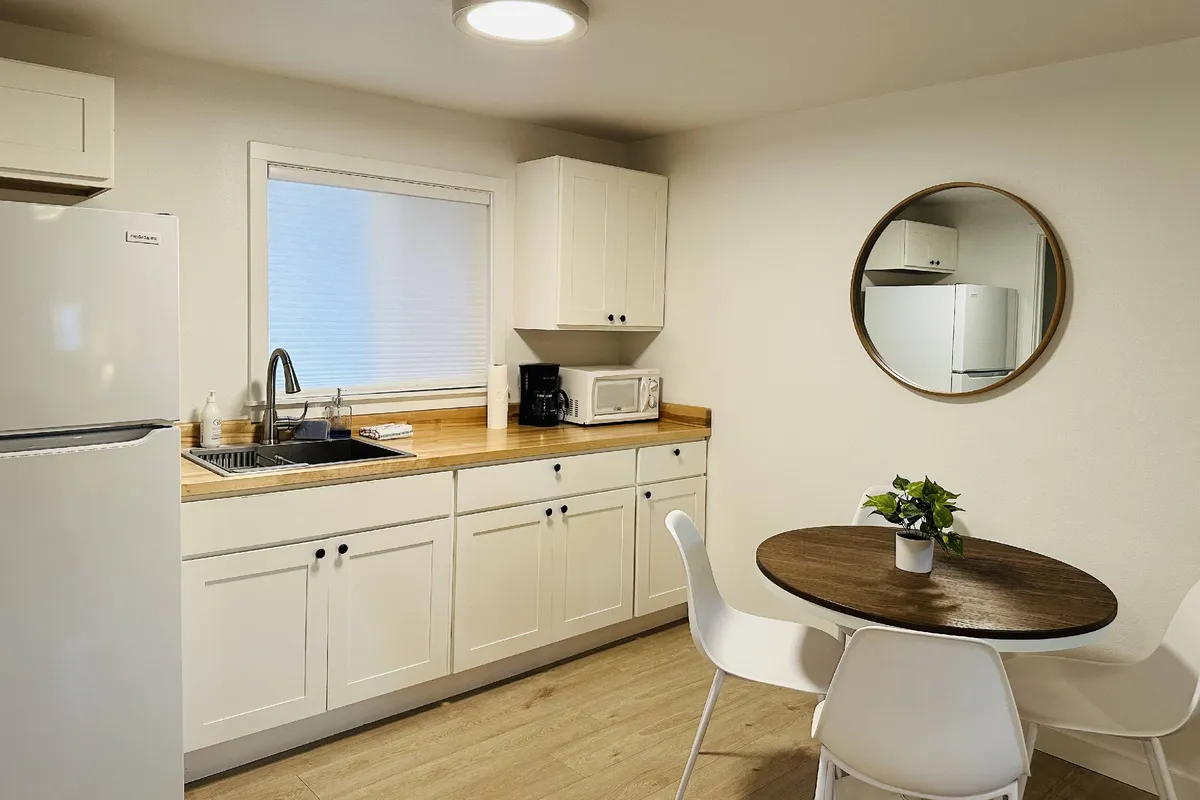 A well-lit kitchenette with a kitchen sink, round dining table set for two, a wall mirror, and a refrigerator. The space radiates warmth and practicality, ideal for preparing meals or enjoying a casual breakfast.