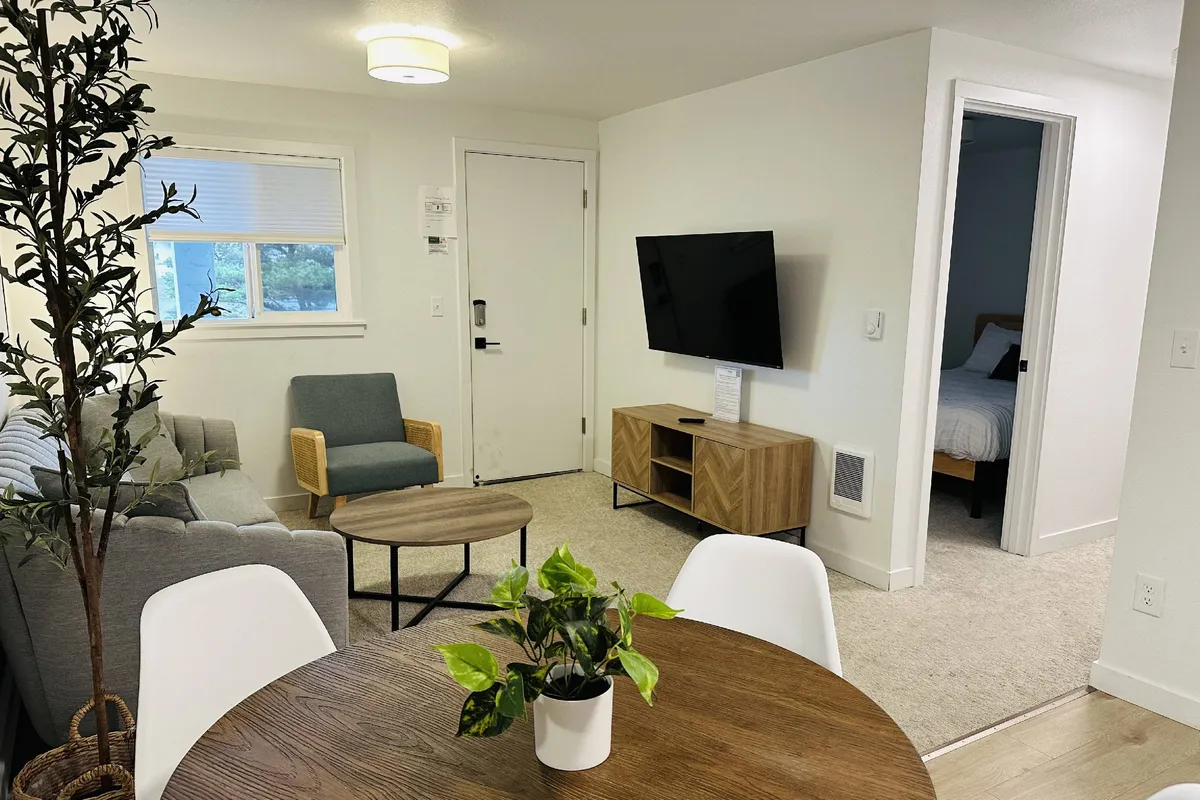 The image showcases a bright, contemporary living and dining area with a comfortable sofa facing a sleek TV, a modern coffee table, and a dining table perfect for sharing meals. The space blends a cozy ambiance with stylish touches, inviting guests to relax, watch movies, or enjoy memorable dinners together after a day at the beach.