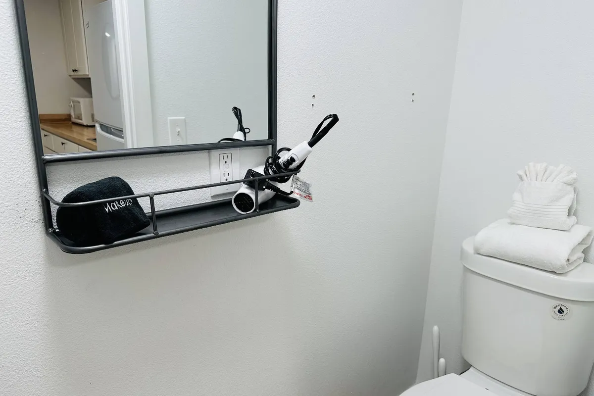 A tidy, stylish bathroom area featuring a wall-mounted mirror, a shelf holding a hair dryer and hat, and neatly arranged toiletries on the toilet tank. The space is clean, functional, and thoughtfully organized for guests' daily routines.