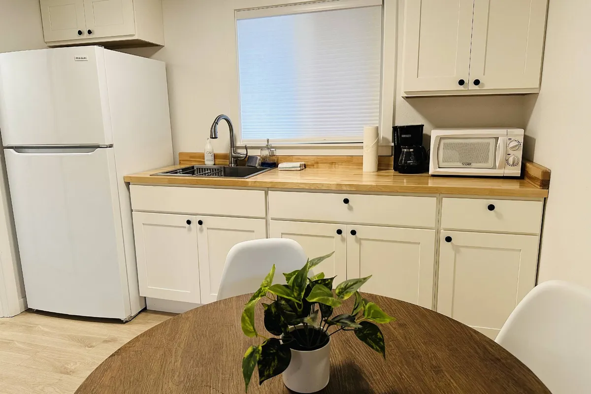 A cozy kitchenette featuring a white refrigerator, kitchen sink, wooden countertop, coffee maker, microwave, and a touch of greenery provided by a small plant. A round dining table is nearby, creating a welcoming spot to share meals or prepare snacks.