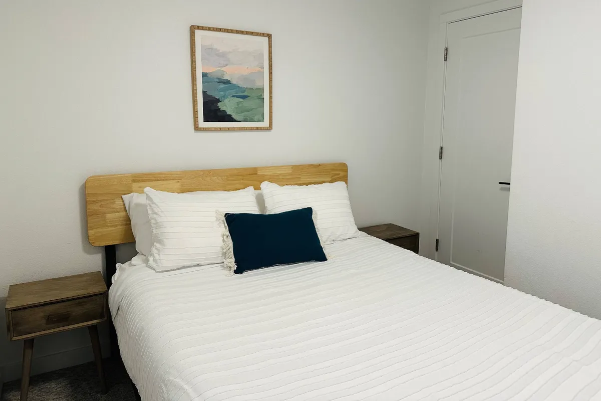 A serene bedroom scene featuring a bed with white textured bedding, a blue accent pillow, and framed artwork on the wall. Soft natural light fills the space, highlighting the tranquil and inviting atmosphere ideal for restful sleep.